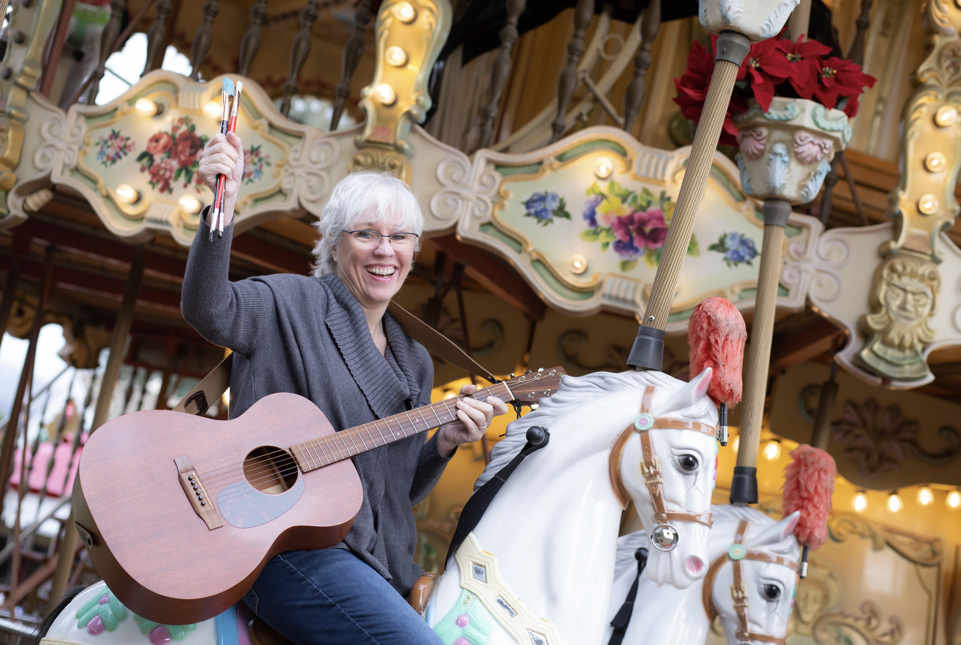 Woman with guitar and paint brushes seated on carousel horse
