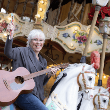 Woman with guitar and paint brushes seated on carousel horse